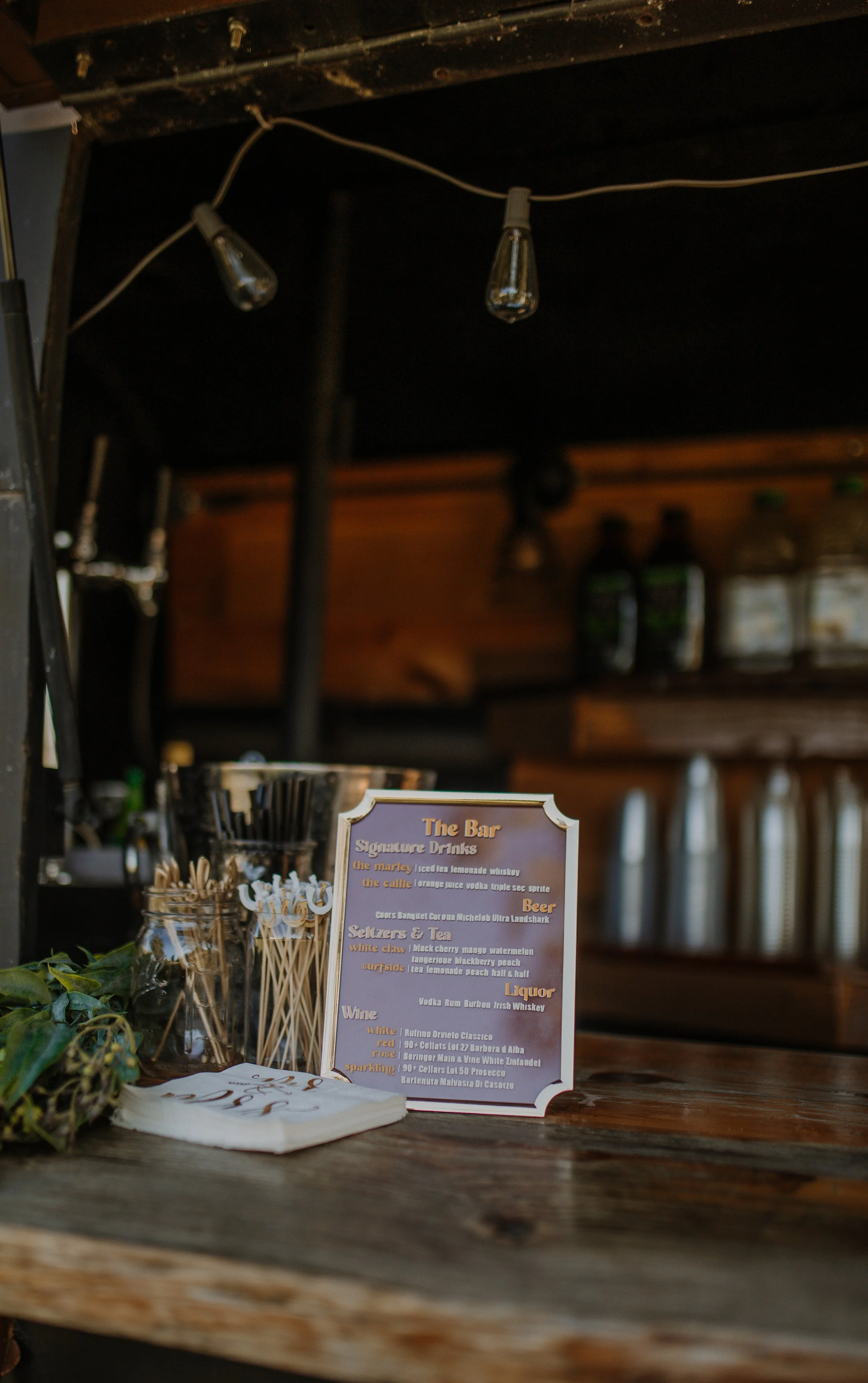 Bar menu on a wooden counter with drink options and a jar of stir sticks, illuminated by hanging string lights, inside a rustic bar.