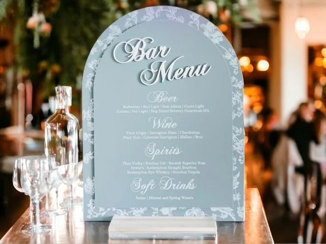 Elegant bar menu sign displaying categories for beer, wine, spirits, and soft drinks in a stylish font with a decorative border, placed on a wooden bar counter with glasses and a bottle nearby.