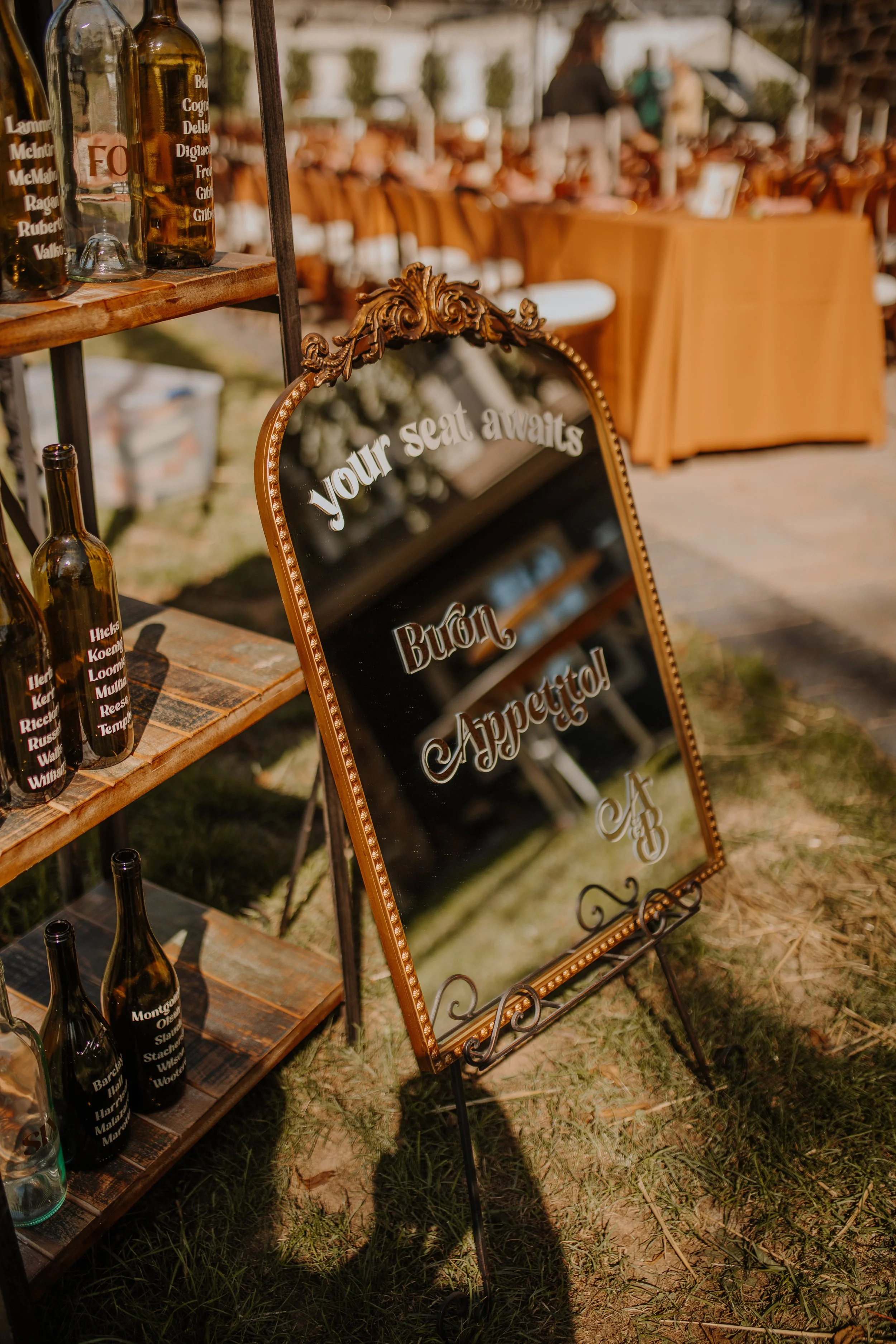 A decorative mirror sign at an outdoor event with the words 'your seat awaits' and 'burn', 'Appreciated', and initials 'A B'. Surrounding it are shelves with glass bottles with printed words on them, and in the background, there are tables and chairs