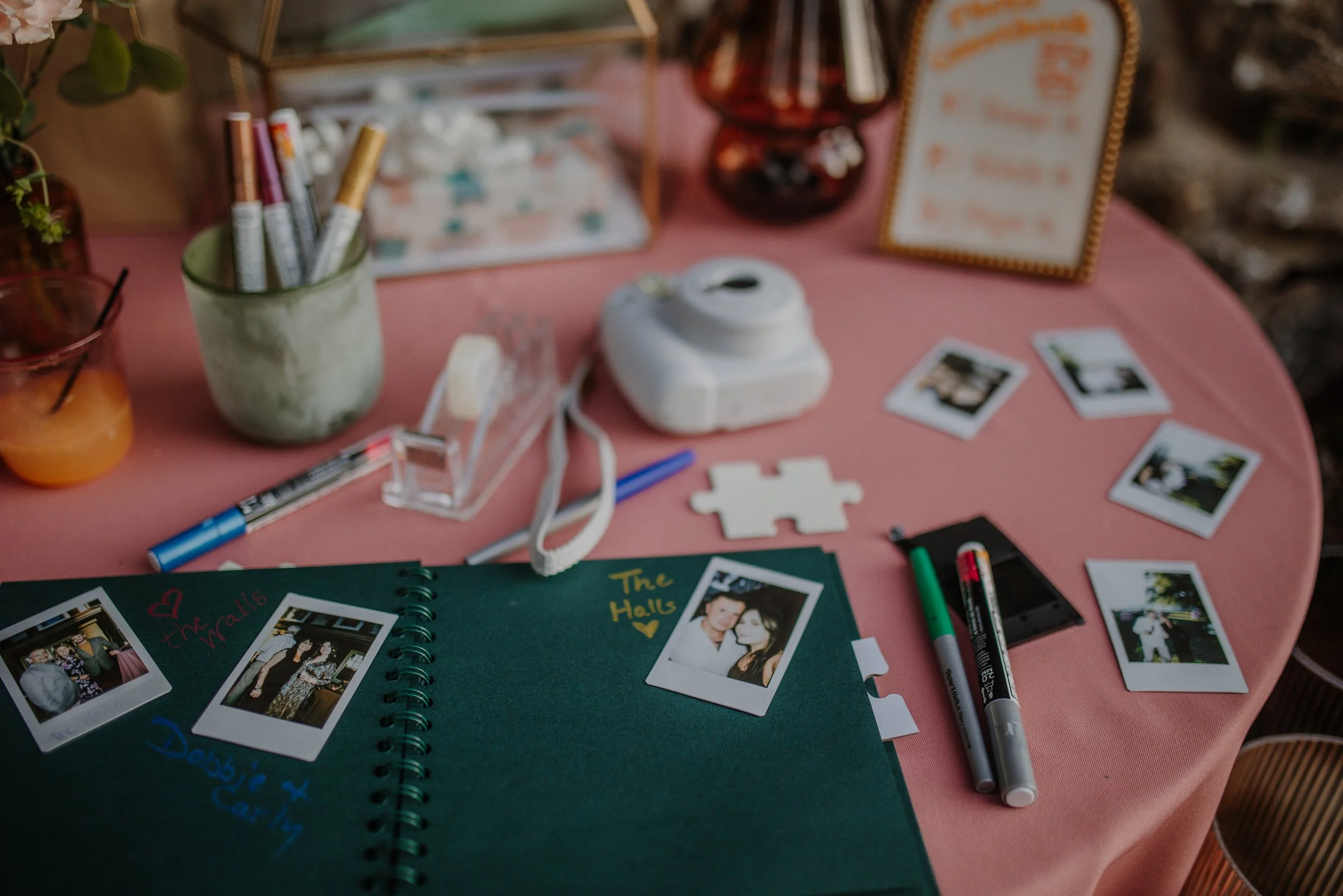 A pink tablecloth on a round table with Polaroid photos, a photo album, and various writing supplies including markers, pens, and tape. There are decorative items such as plants, a framed picture, and jars on the table.