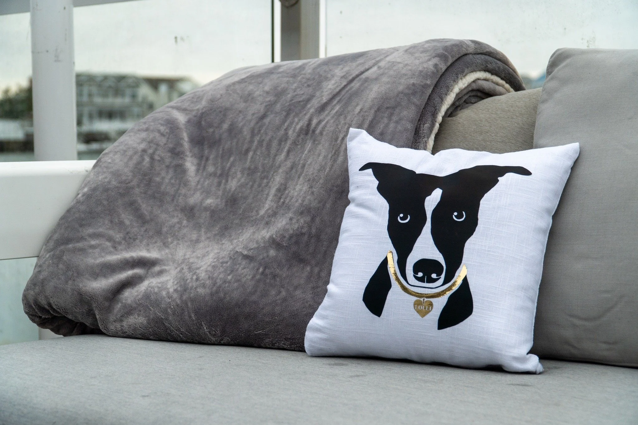 Decorative throw pillow with a black and white dog illustration and a gold necklace with a heart-shaped tag, placed on a beige outdoor sofa with a gray blanket and another gray cushion, overlooking a balcony with water and buildings in the background