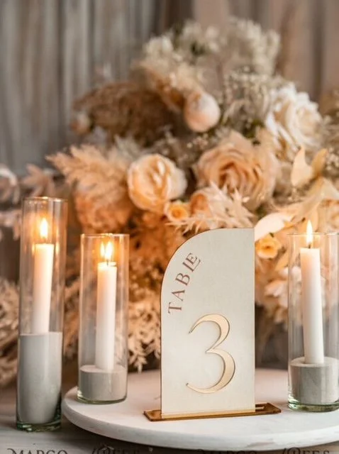 Wedding table centerpiece with cream and blush flowers, lit candles in glass holders, and a table number card marked '3'.