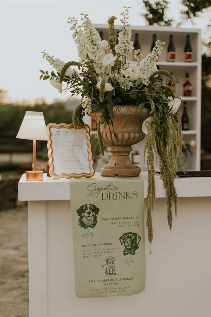 A floral arrangement in an ornate vase, a framed menu, a small white table lamp, and a drink sign with dog-themed drink names at an outdoor event.