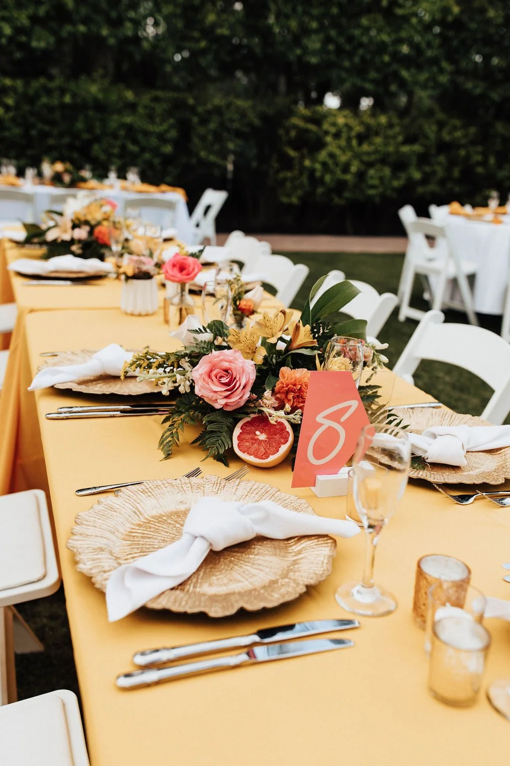 A long outdoor table decorated for a special event with floral centerpieces, grapefruit halves, table settings including napkins, plates, silverware, and glasses, and a pink table card with the number eight.