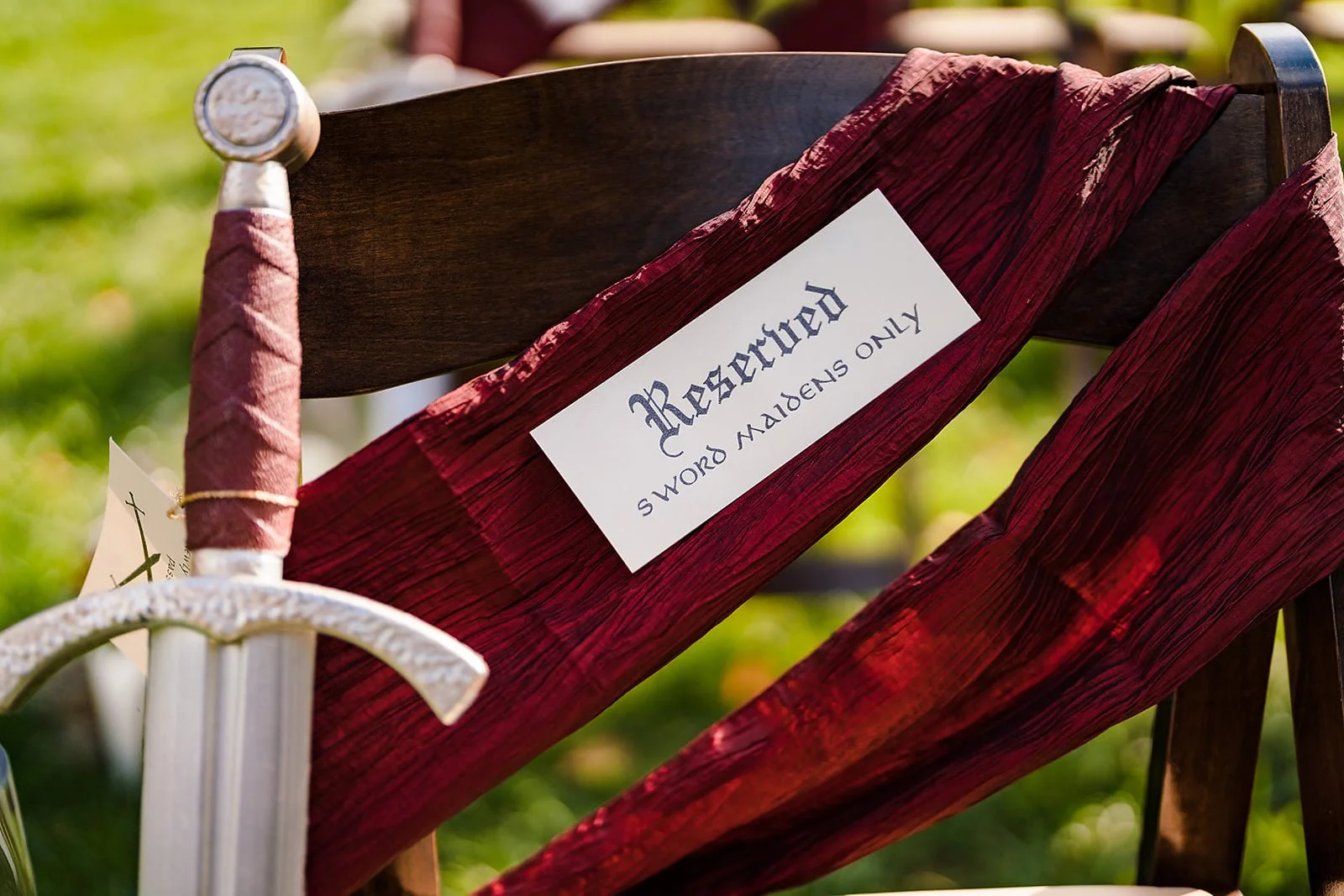 A wooden chair with a red fabric sash and a sign that reads 'Reserved' and 'Swords Only. Please' attached to the back.