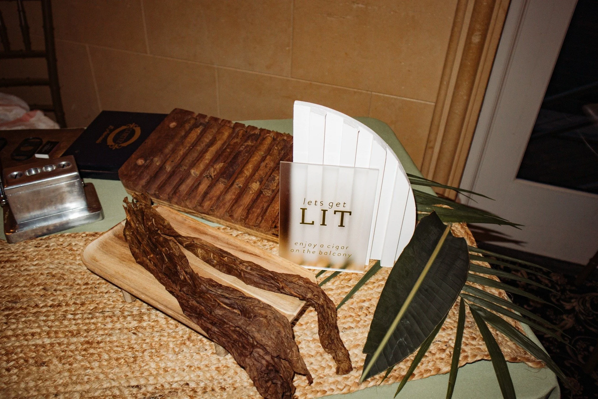 A wooden table displaying cigar-related items including a tray of cigars, a sign that says "let's get LIT" with the subtitle "enjoy a cigar on the balcony," and a dried cigar leaf. The setup is on a woven mat with some green leaves for decoration.