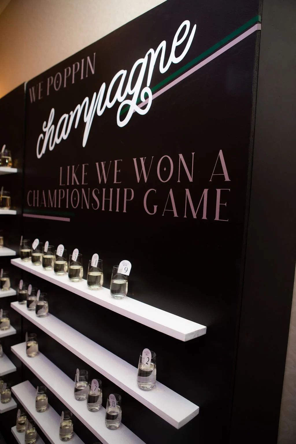 Display of wine glasses with numbered tags under a sign that reads 'We Poppin Champagne like we won a championship game' in a wine store or tasting room.