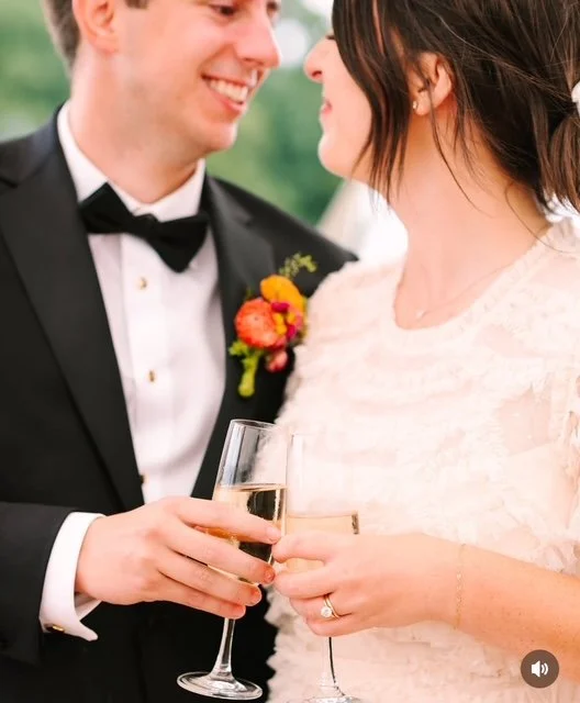 A bride and groom smiling at each other, holding glasses of champagne, during their wedding celebration. The groom is wearing a black tuxedo with a bow tie and a boutonniere. The bride is wearing a white wedding dress with lace detail and has a gold
