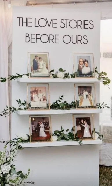 A white display stand with three shelves decorated with green leaves and white flowers, showing framed wedding photos. A sign above reads "The Love Stories Before Ours."