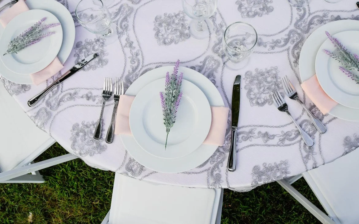Table setting with a pink tablecloth, white plates, silverware, empty glasses, and a lavender sprig on each plate. Two white chairs are partially visible, and the table is outdoors on grass.