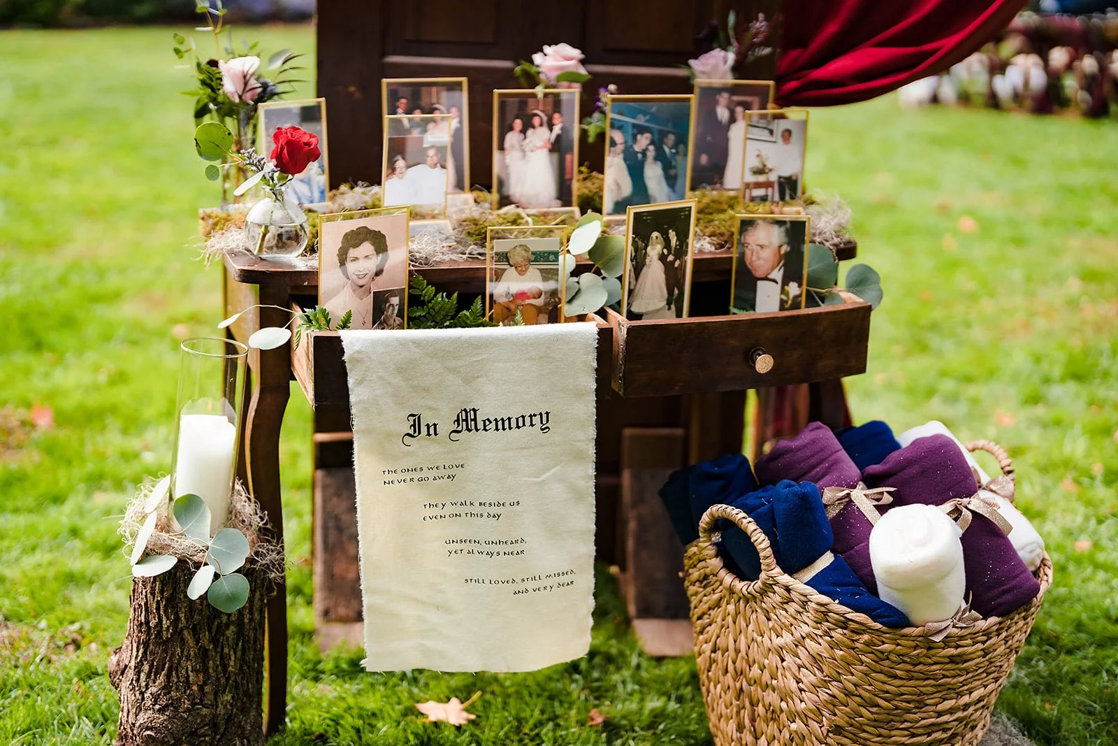 A memorial display with framed photographs, a white cloth with a memorial message, a basket of folded clothes, and candles arranged outdoors on green grass.