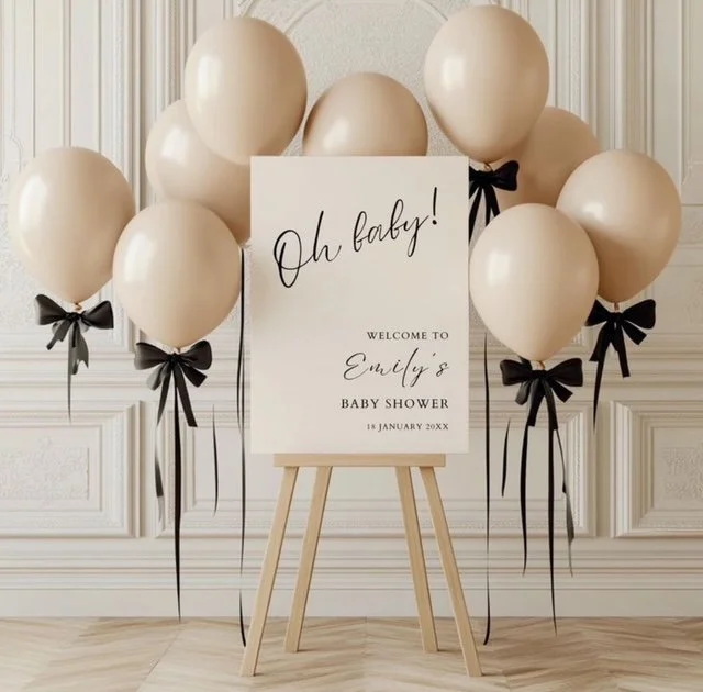 Celebratory white balloons with black ribbons arranged around a welcome sign for Emily's baby shower, dated January 18, 20XX, against a white paneled wall.
