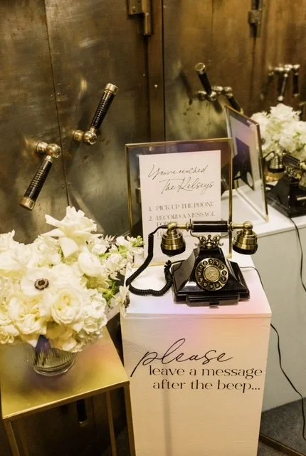 Vintage black rotary phone on a pink donation box with a message to leave a message after the beep, surrounded by white flowers and informational signs in the background.