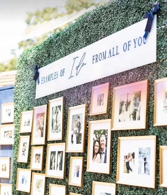 Photo of a display board with framed photos and a sign that reads 'Examples of Love from All of You' at a wedding or celebration.