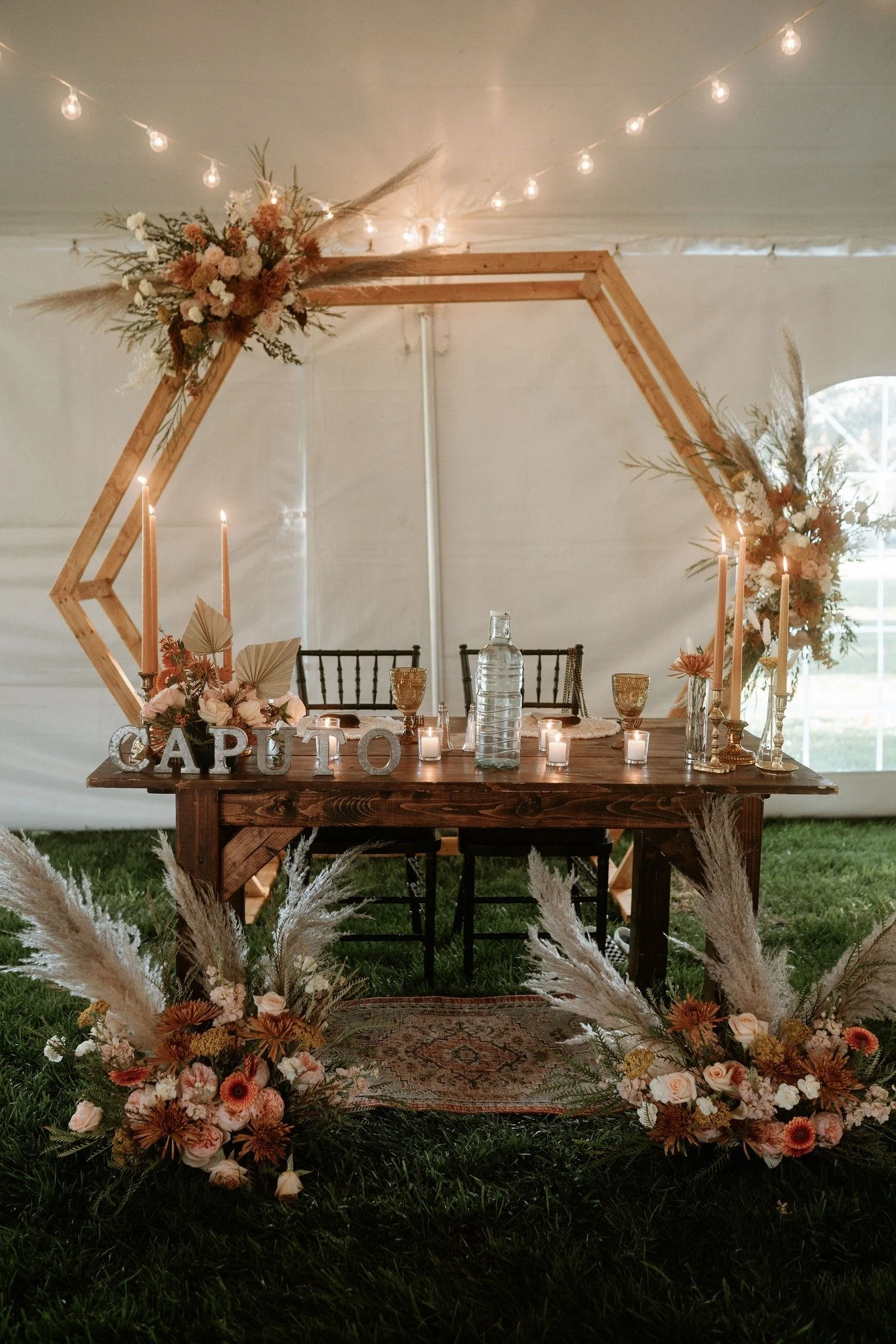 Indoor wedding reception with a wooden table decorated with candles, flowers, and a water bottle, set in front of a geometric wooden arch adorned with floral arrangements, string lights overhead, and a rug on the grass floor.
