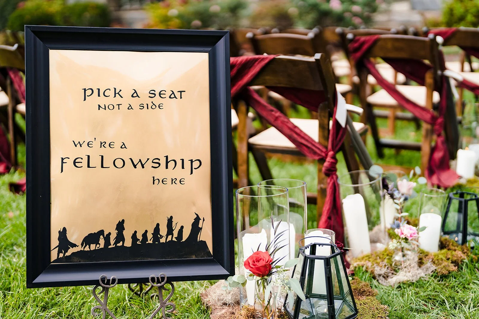 Wedding sign that says 'Pick a seat, not a side, we're a fellowship here,' placed on a stand at an outdoor ceremony with wooden chairs tied with red ribbons, candles, and floral decorations.