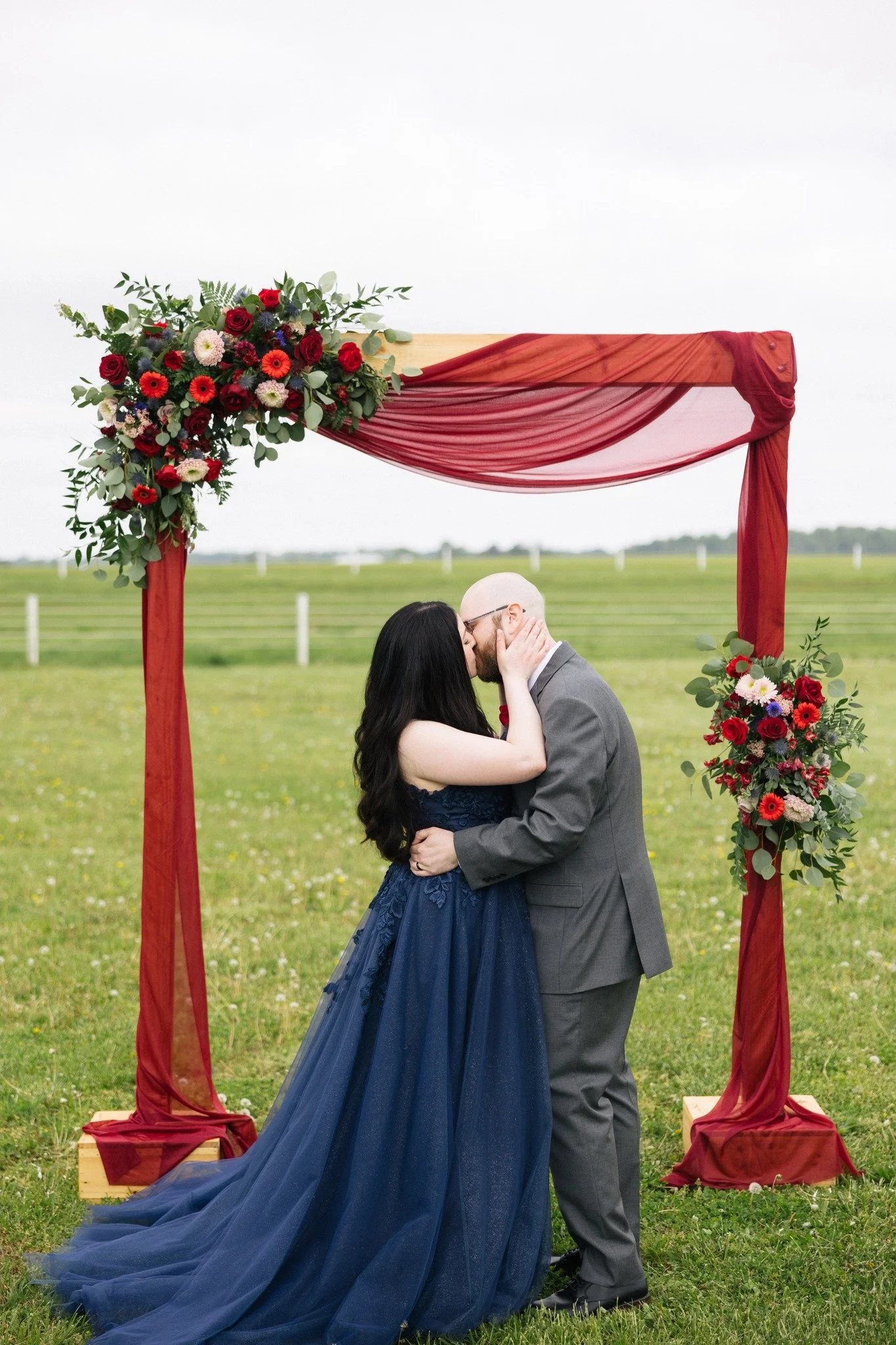 A couple sharing a kiss under a wedding arch decorated with red and pink flowers and red fabric, set in an outdoor grassy field.
