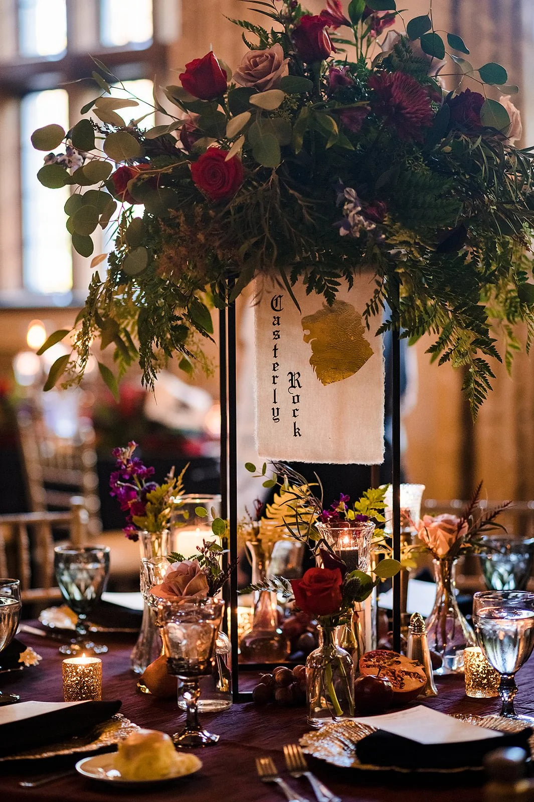 A wedding table centerpiece with a tall floral arrangement, candles, and water glasses, featuring a sign reading 'Casterly Rock' with a gold leaf design.