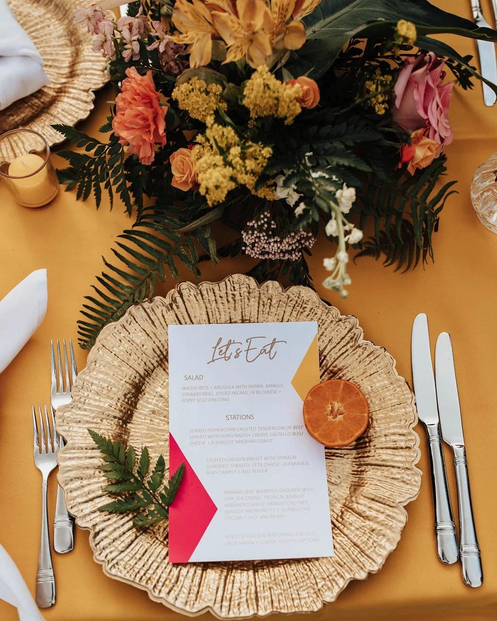 A table setting with a gold decorative charger plate, a white menu with gold and red accents, a sprig of green fern, and an orange slice. There are silver utensils and a floral centerpiece with pink, yellow, and white flowers and greenery. A small gl