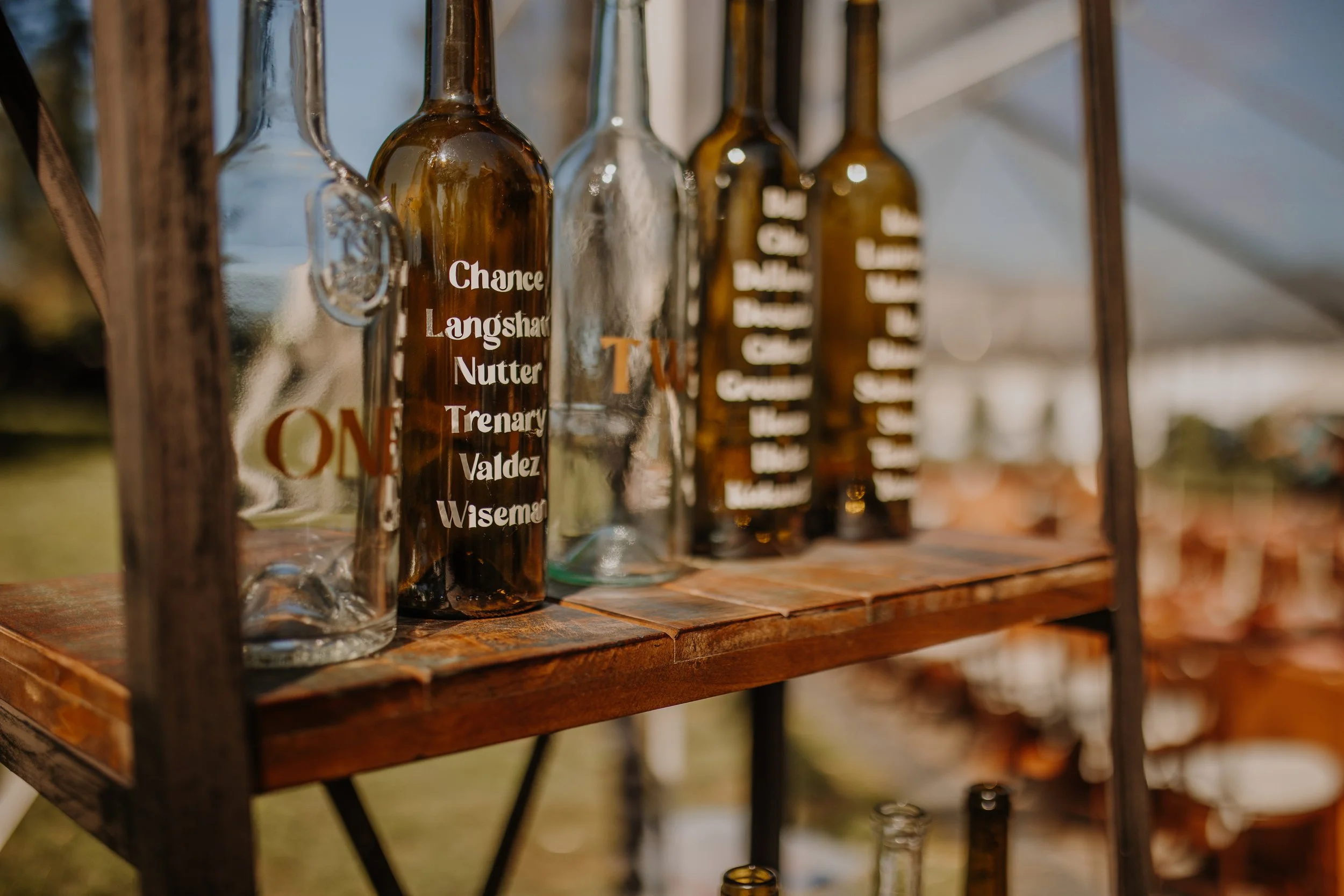 Several glass bottles with painted names, displayed on a wooden shelf at an outdoor event.