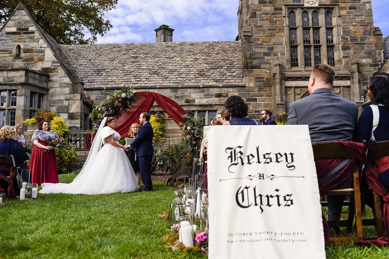 A wedding ceremony taking place outside a stone church, with a bride and groom exchanging vows under a floral arch. Guests are seated, and a sign with the names 'Kelsey and Chris' along with the date October twenty-fourth, two thousand and twenty-one