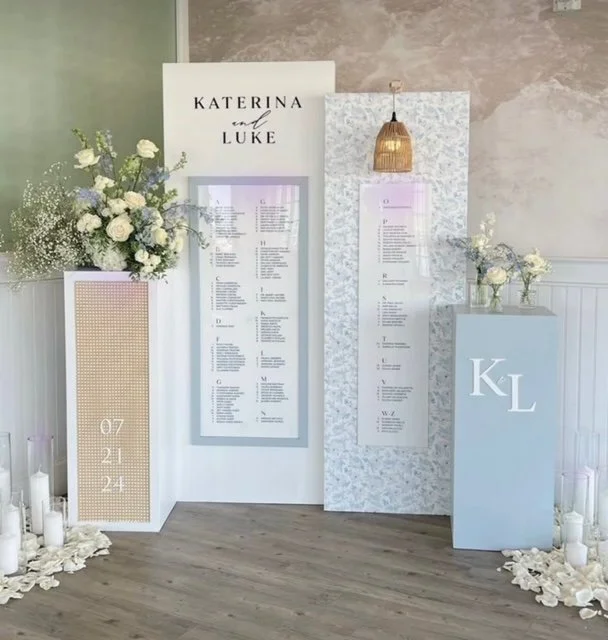 Wedding or event seating chart display with floral arrangements, candles, and decorative panels. Names 'Katerina and Luke' are at the top, with date '07.21.24' on a side panel and initials 'KL' on a pedestal.