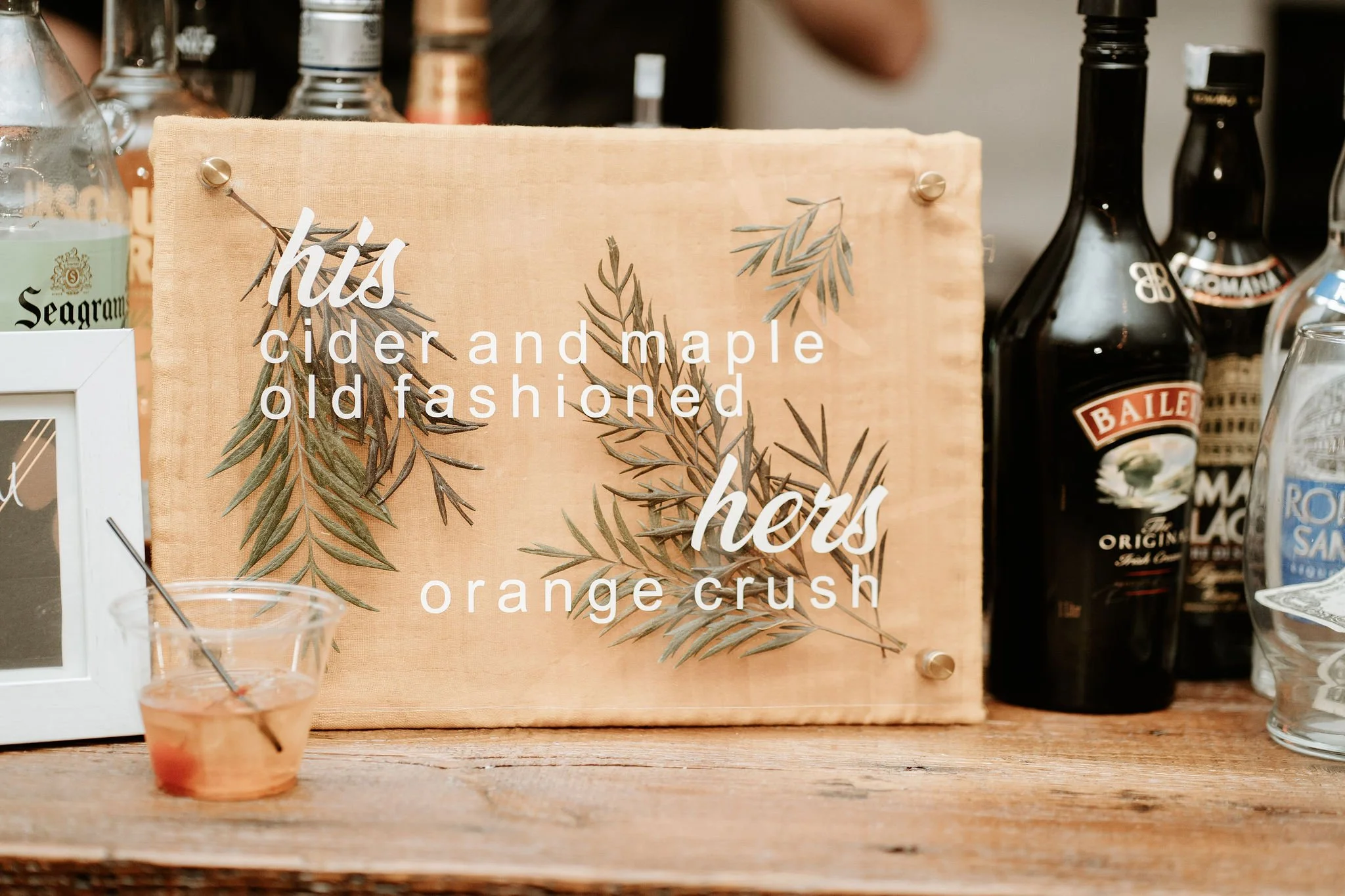 A decorative wooden sign with dried green leaves and white text that reads 'his cider and maple old fashioned hers orange crush' displayed on a wooden surface, surrounded by bottles and glasses.