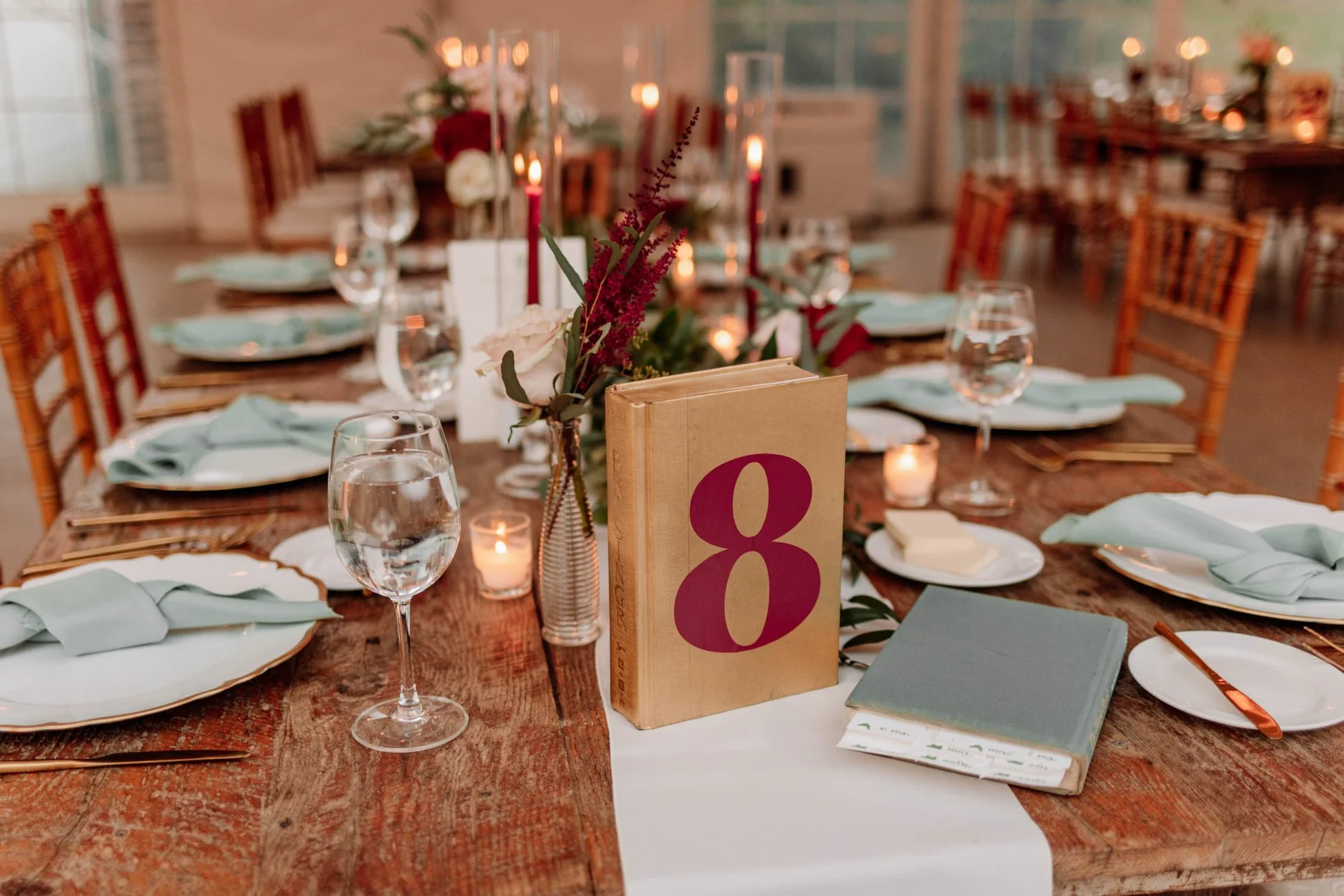 Table set for a formal event with a rustic wooden table, cloth napkins, glassware, plates, the number 8 on a gold stand, a small bouquet of flowers, candles, and additional chairs and tables in the background.