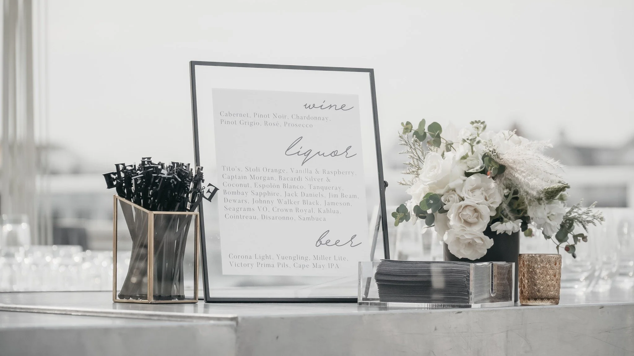 A table setup with a framed menu listing wine, liquor, and beer options, a container holding black pens, a stack of paper napkins, and a vase with white roses and green foliage.