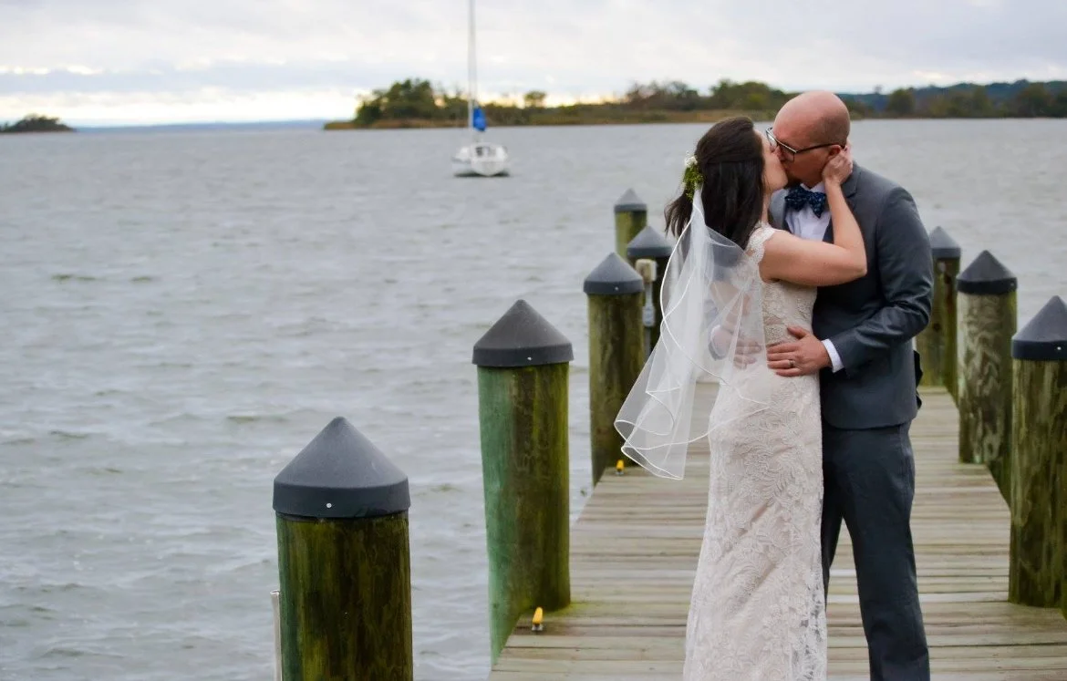 A bride and groom kissing on a wooden dock by a body of water, with a sailboat in the background.
