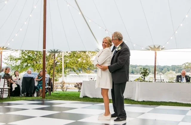 A bride and groom share a dance under a white tent at an outdoor wedding reception, with guests seated at tables in the background.