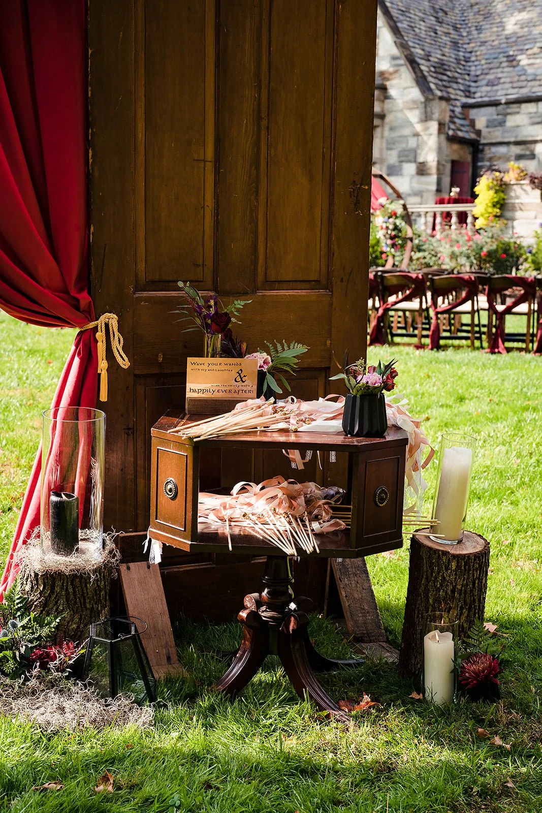 Decorative outdoor wedding setup with candles, flowers, and ribbons on a wooden table and harvest-style logs on a lawn, with chairs and garden in the background.