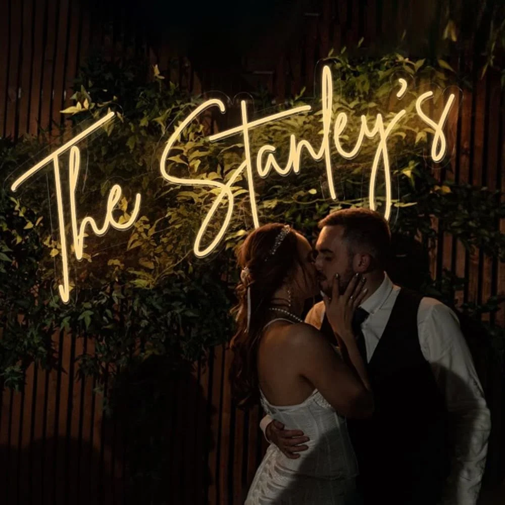 A couple kissing in front of a neon sign that says 'The Stantys' with greenery behind them.
