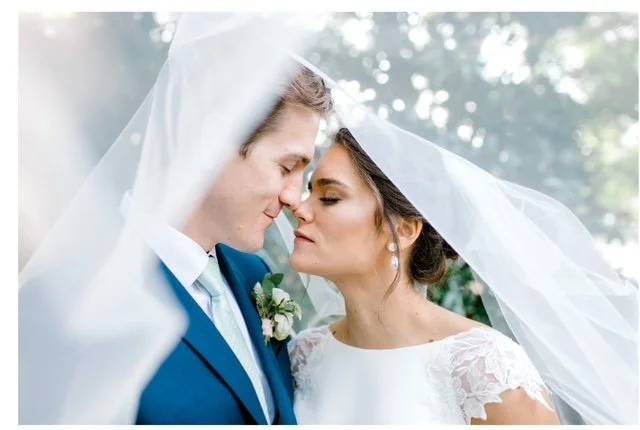 Bride and groom with their foreheads touching under a wedding veil in a garden setting.
