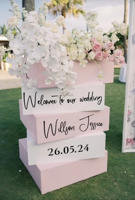 A wedding welcome sign with a floral arrangement on top, displaying the text "Welcome to our wedding Willson Jessica 26.05.24" on pink and white blocks.
