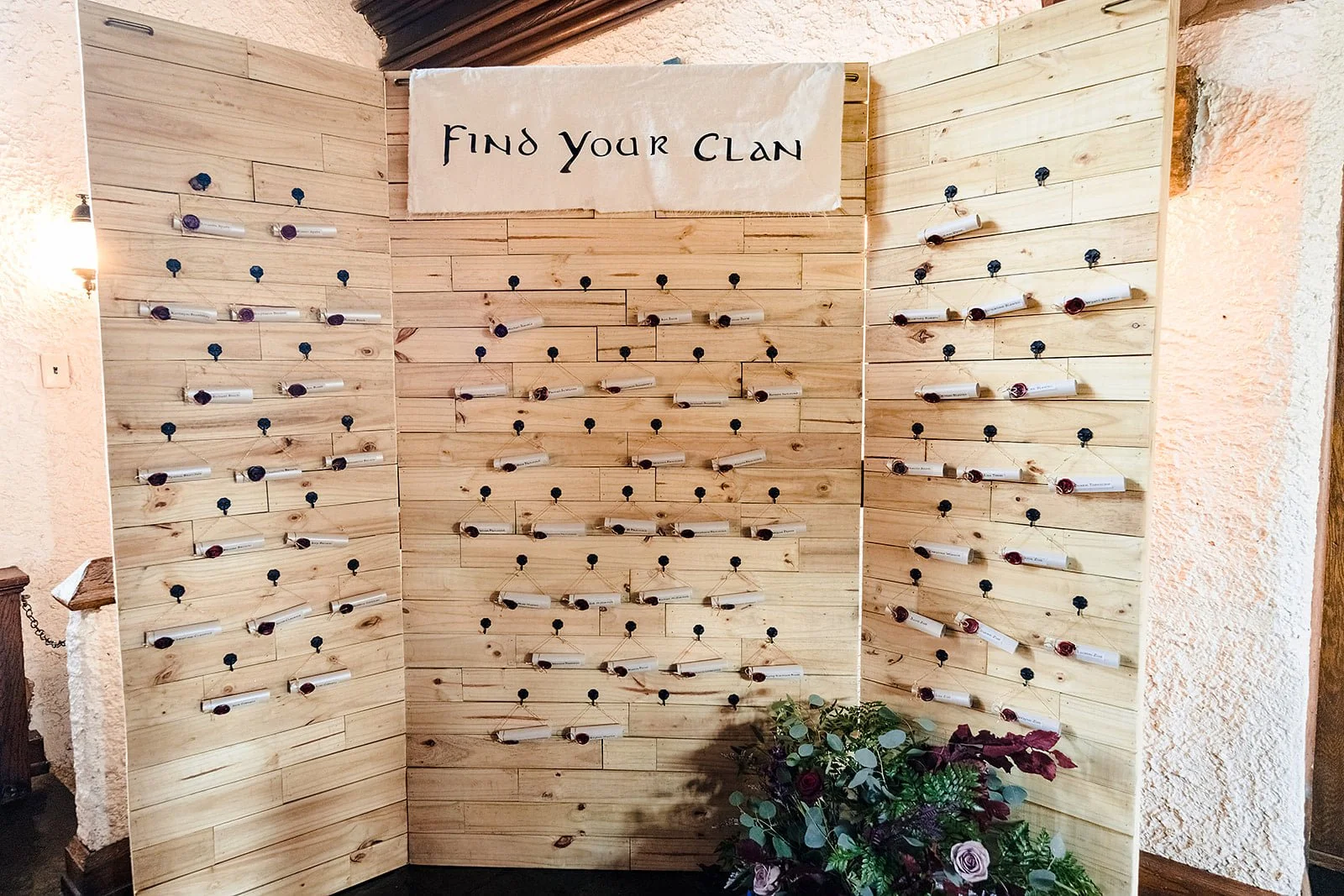 A wooden wall display with small paper scrolls attached by strings and pins, labeled with different names and colors of wine bottles, with a sign above that says 'Find Your Clan', and a floral arrangement at the bottom.