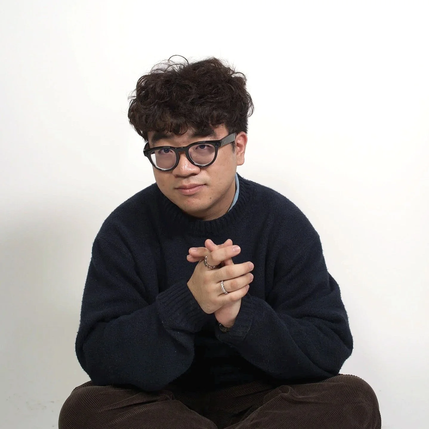 A young man with curly dark hair and glasses, wearing a dark sweater and rings on his fingers, sitting cross-legged against a plain white wall, with hands clasped in front of him.