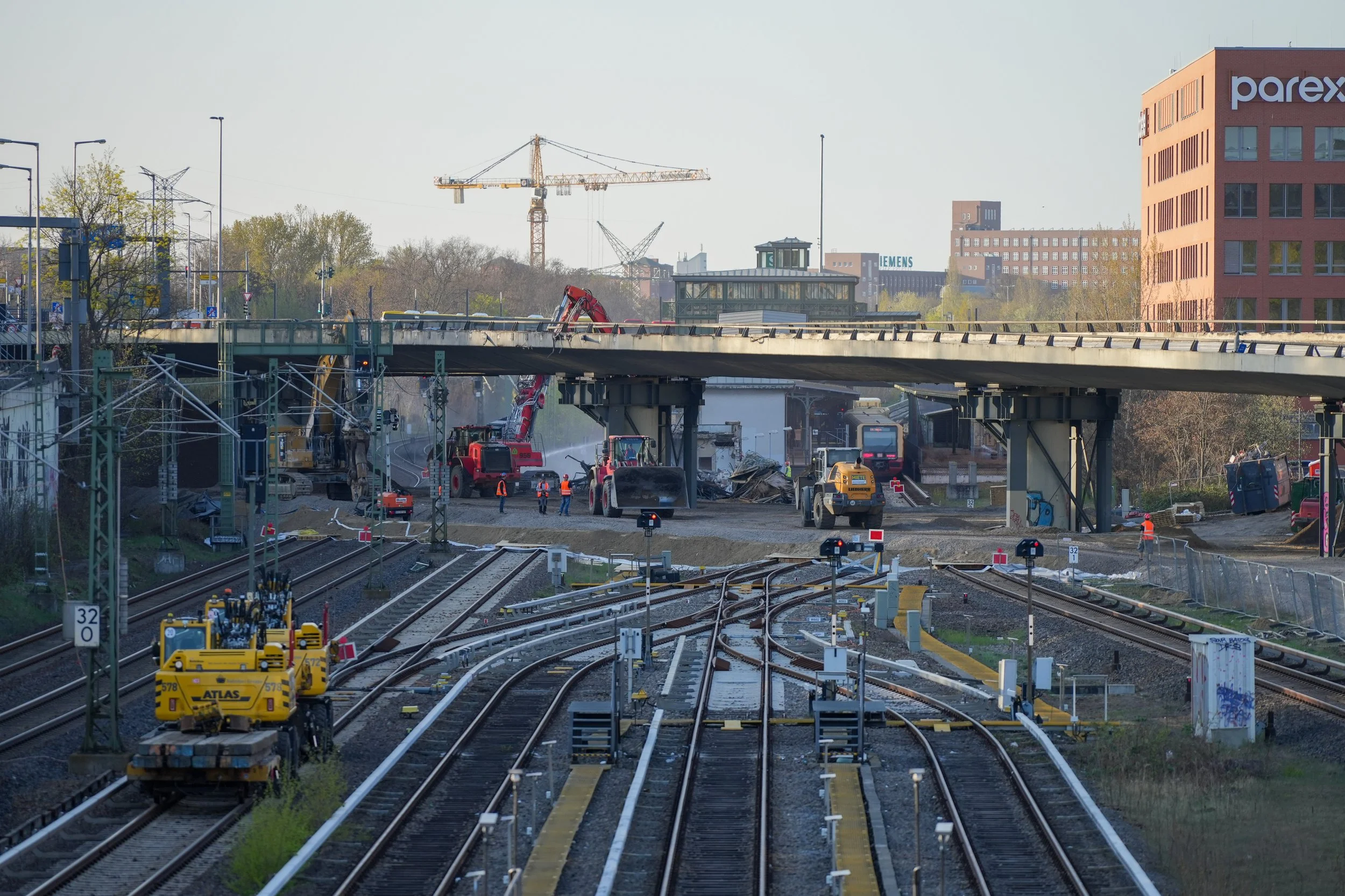 Bauarbeiten an einer Eisenbahnüberführung mit Baustellenfahrzeugen und Arbeitern auf einer Baustelle. Im Hintergrund sind Kräne und Bürogebäude sichtbar.