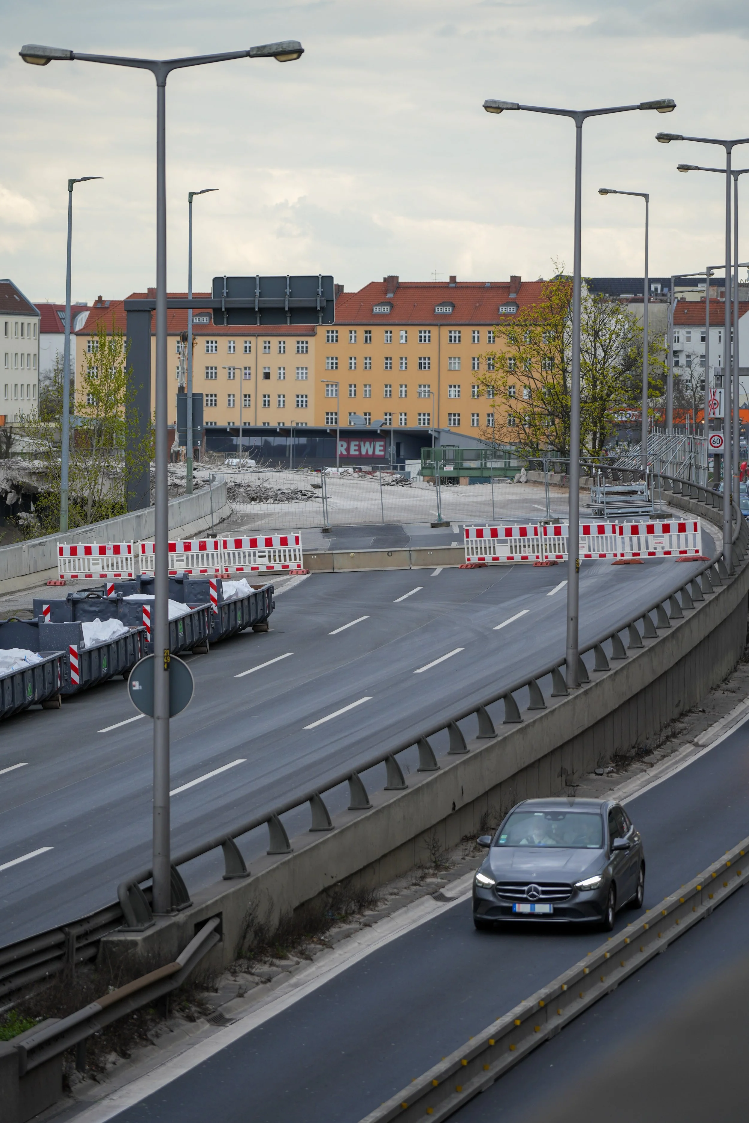 Stadtstraße mit Baustelle und Absperrungen, im Hintergrund Gebäude mit REWE-Logo, ein fahrendes Auto auf einer Spur.