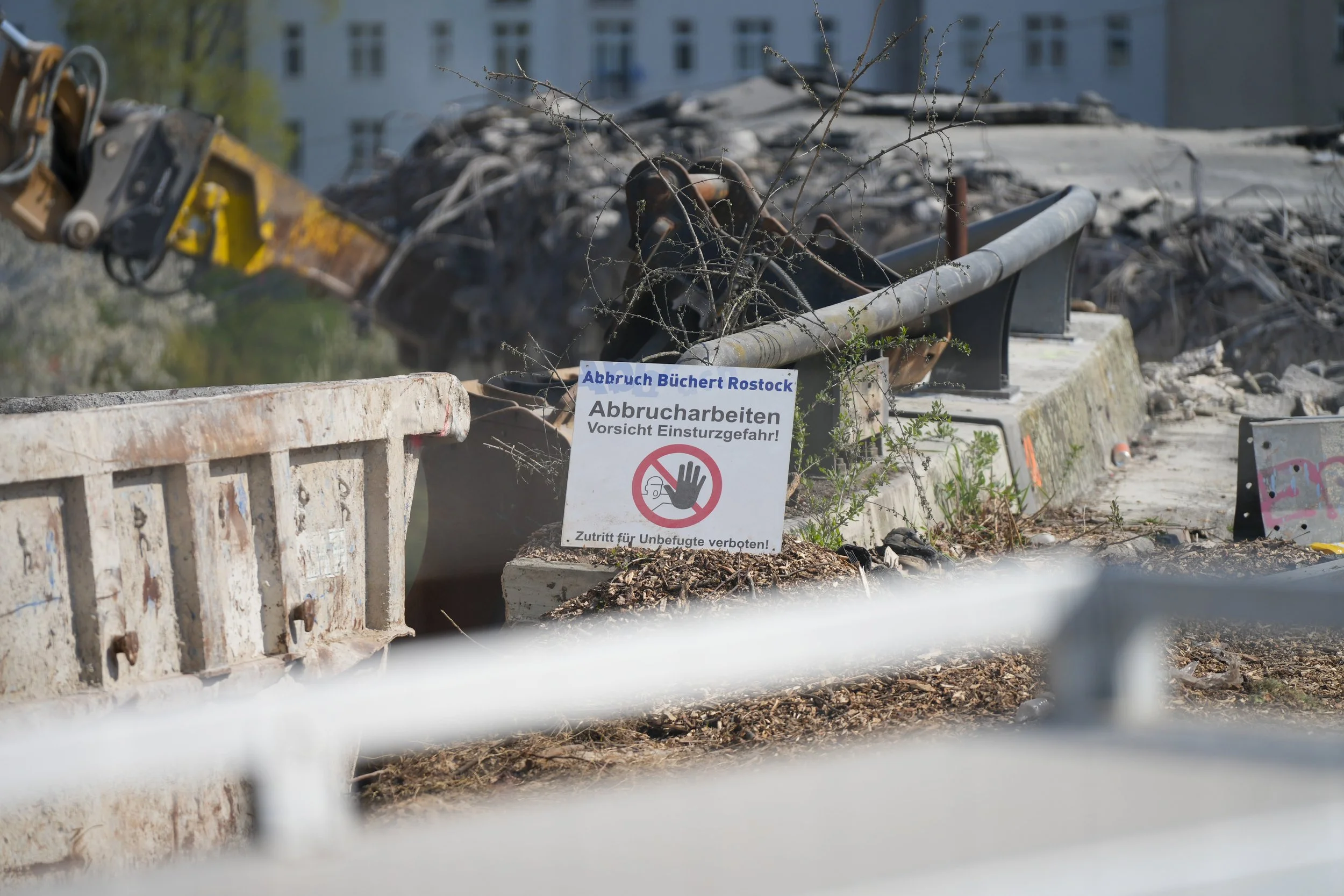 Abbruchstelle mit Schild "Abbruch Büchert Rostock Abbrucharbeiten Vorsicht Einsturzgefahr! Zutritt für Unbefugte verboten!", daneben ein Baggerarm und Schutt.