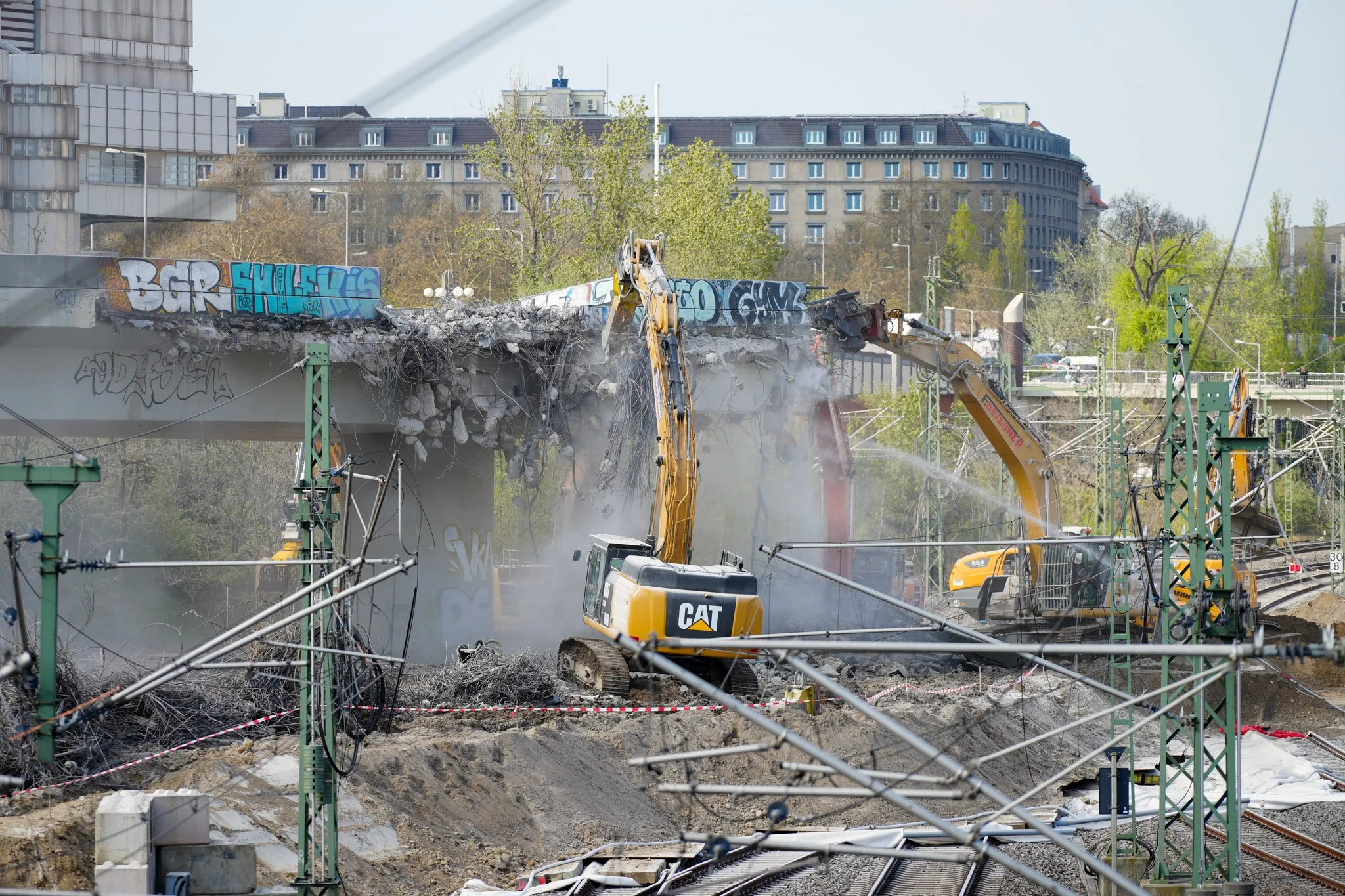 Abriss einer Brücke über Eisenbahnschienen, mit Baggern, Graffiti auf Beton und umliegenden Gebäuden im Hintergrund.