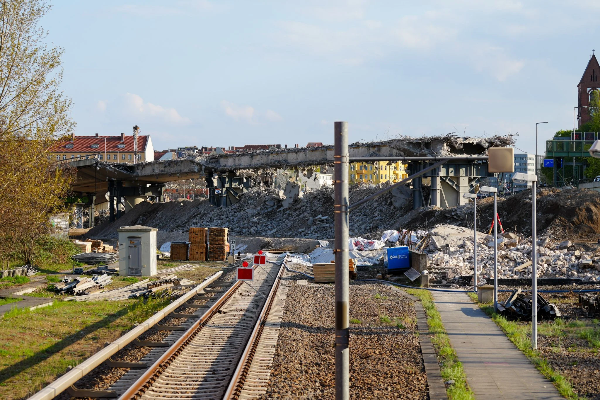 Baustelle mit zerstörter Eisenbahnbrücke, Schutt und Materialien entlang der Gleise, umgeben von städtischer Architektur.