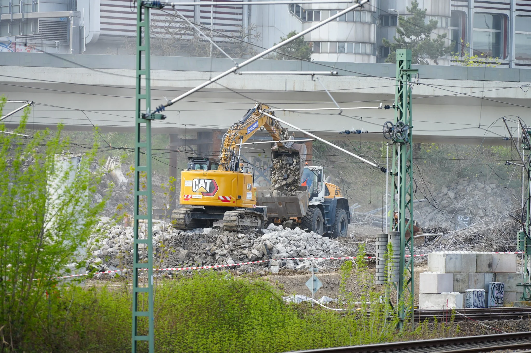 Bauarbeiten an einer Baustelle mit Bagger und Radlader, Schutt und Eisenkonstruktionen im Vordergrund, Eisenbahnschienen und Grünpflanzen im Vordergrund, moderne Gebäude im Hintergrund.