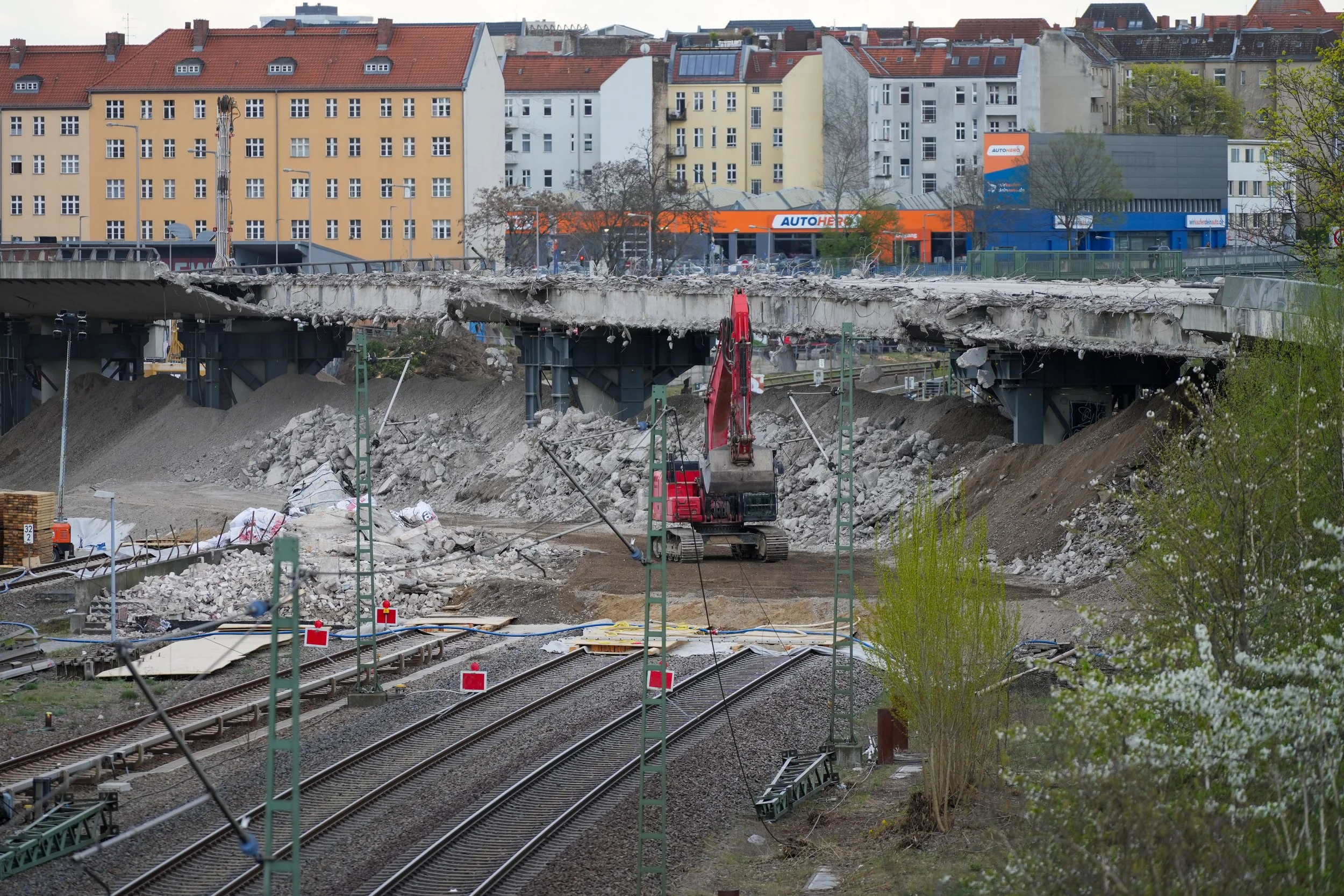 Abbruch einer Brücke über Bahngleisen mit einem Bagger, umgeben von Gebäuden im Hintergrund.