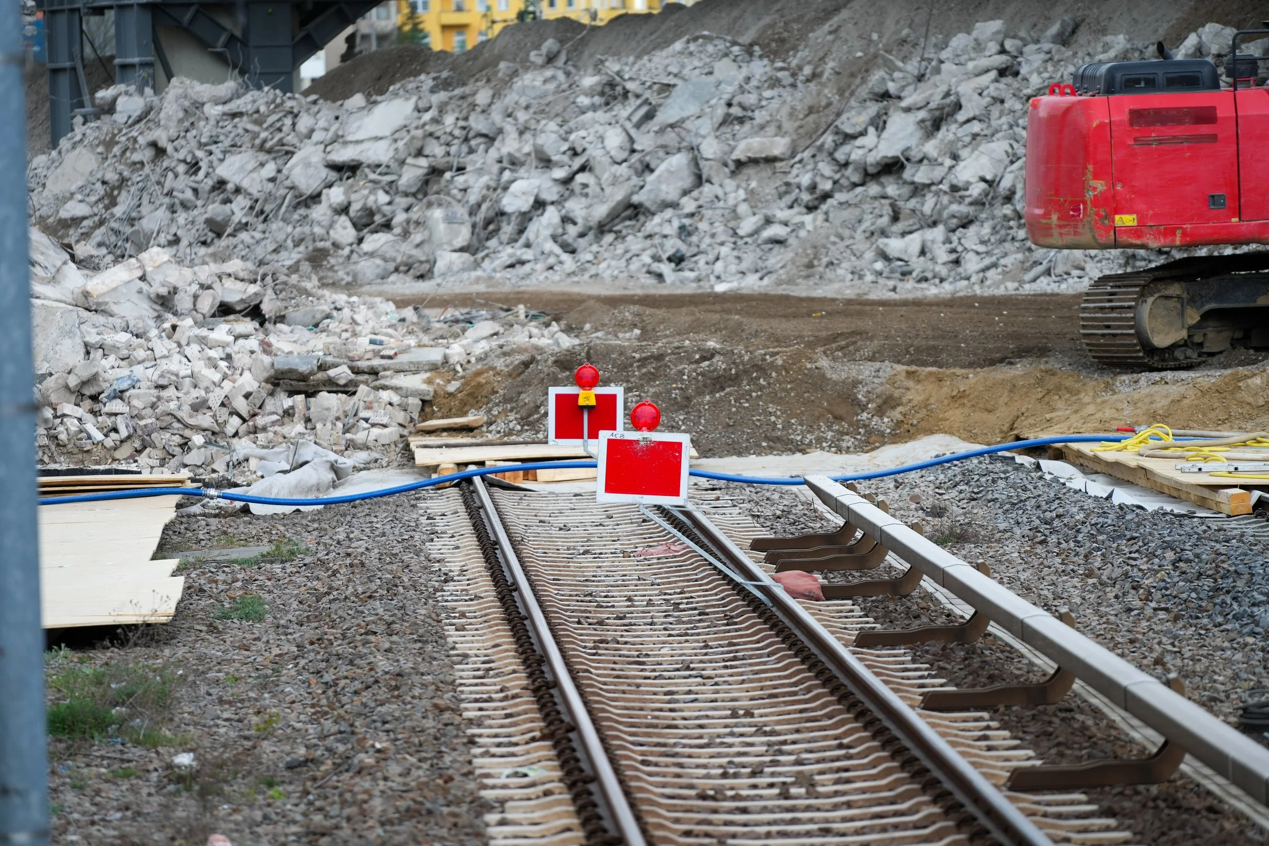 Eine Baustelle mit Eisenbahnschienen, die teilweise entfernt sind, mit Warnschildern und einem roten Bagger im Hintergrund. Im Hintergrund sind Schutthaufen zu sehen.