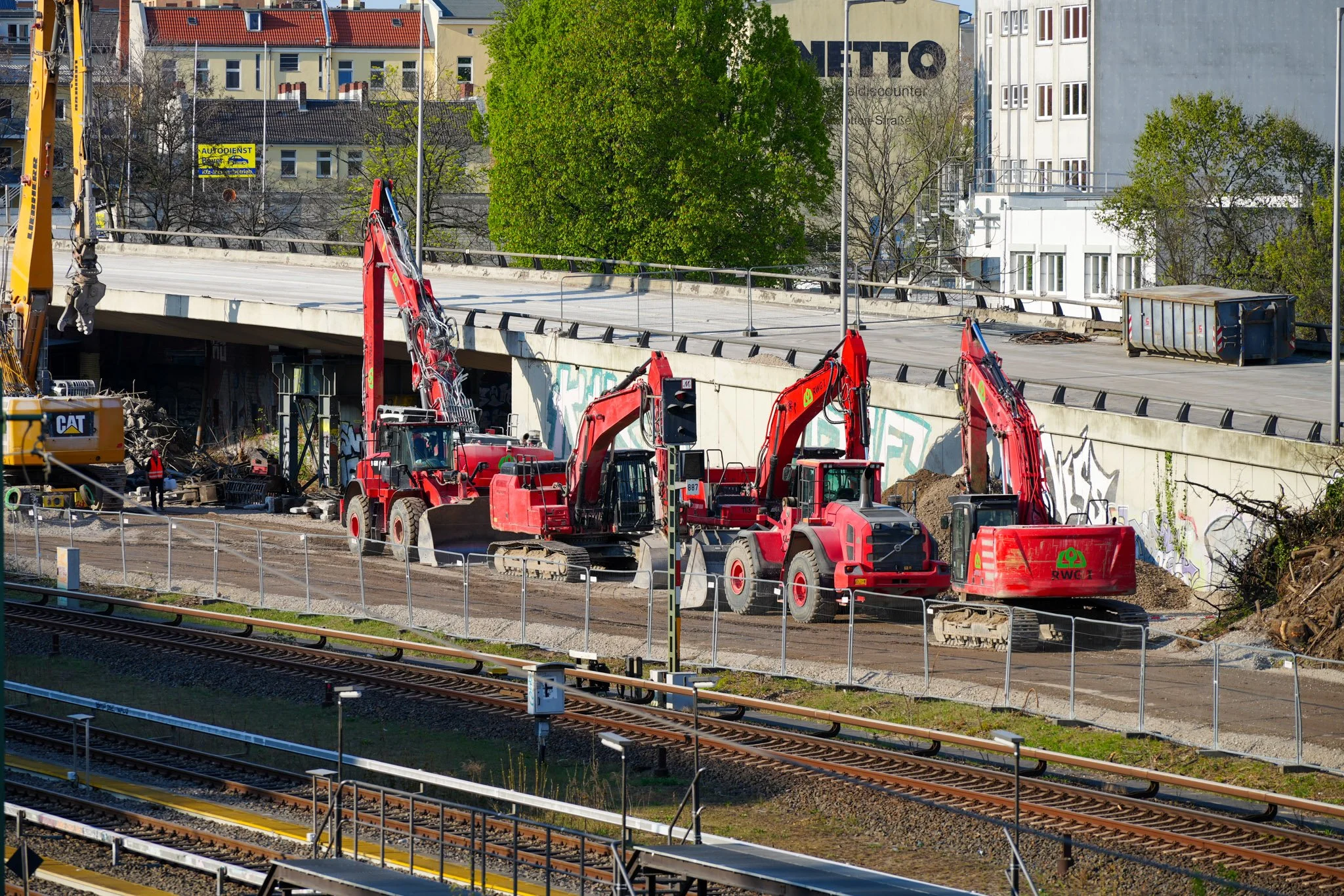 Baustelle mit mehreren roten Baggern und Baumaschinen nahe einer Eisenbahnlinie und einer Brücke. Im Hintergrund sind Gebäude und ein Baum sichtbar.