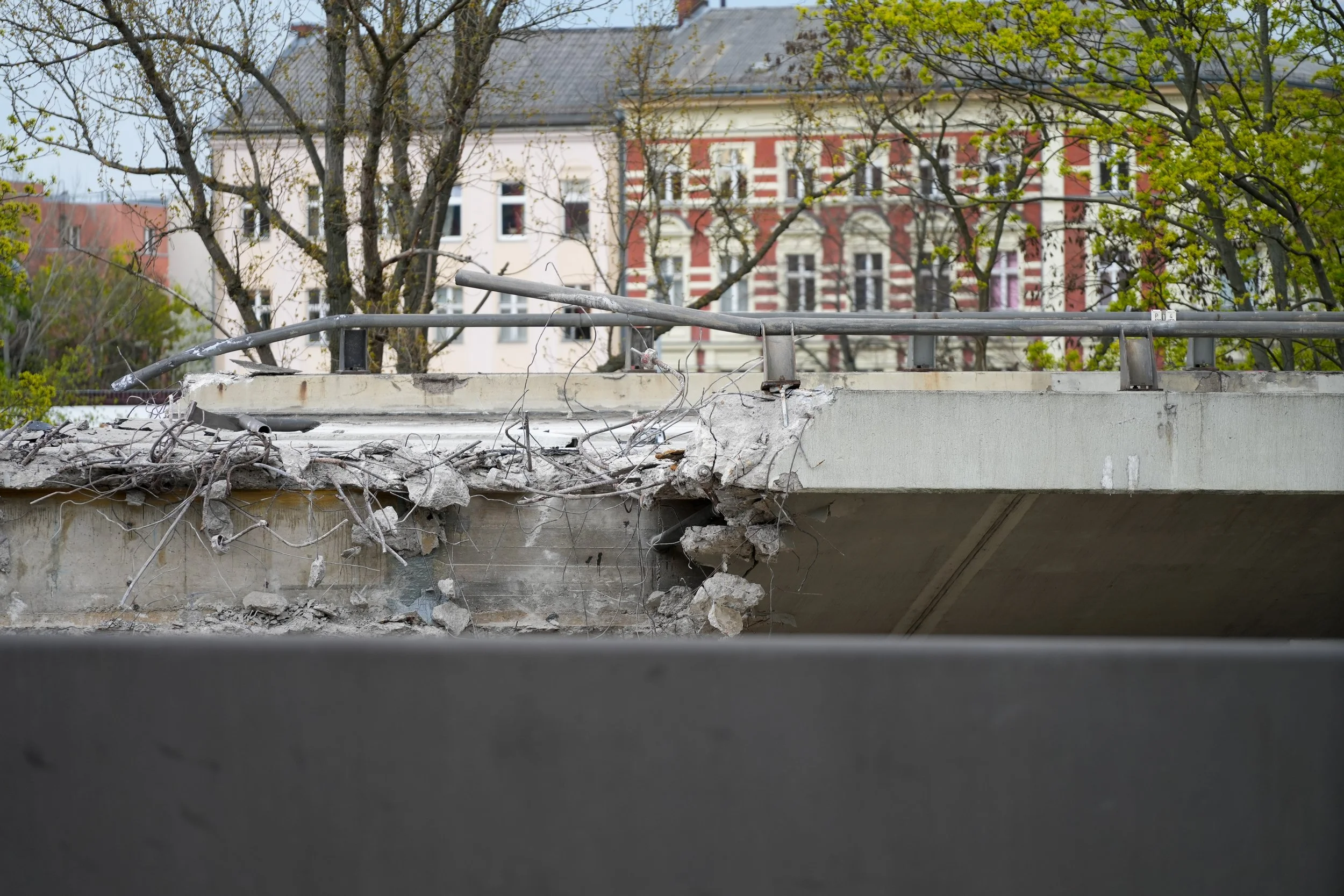 Zerbrochene Brücke mit freiliegenden Stahlstäben und beschädigtem Beton vor einem Altbau im Hintergrund.