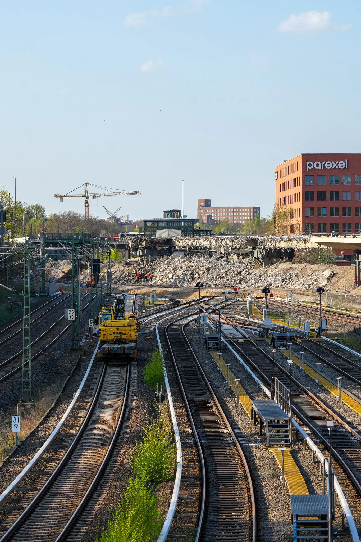 Eisenbahnstrecken mit einem gelben Wartungsfahrzeug, daneben eine Baustelle mit abgerissenen Brückenresten und Bauarbeitern. Im Hintergrund ist ein rotes Gebäude mit der Aufschrift "Parexel" sichtbar.