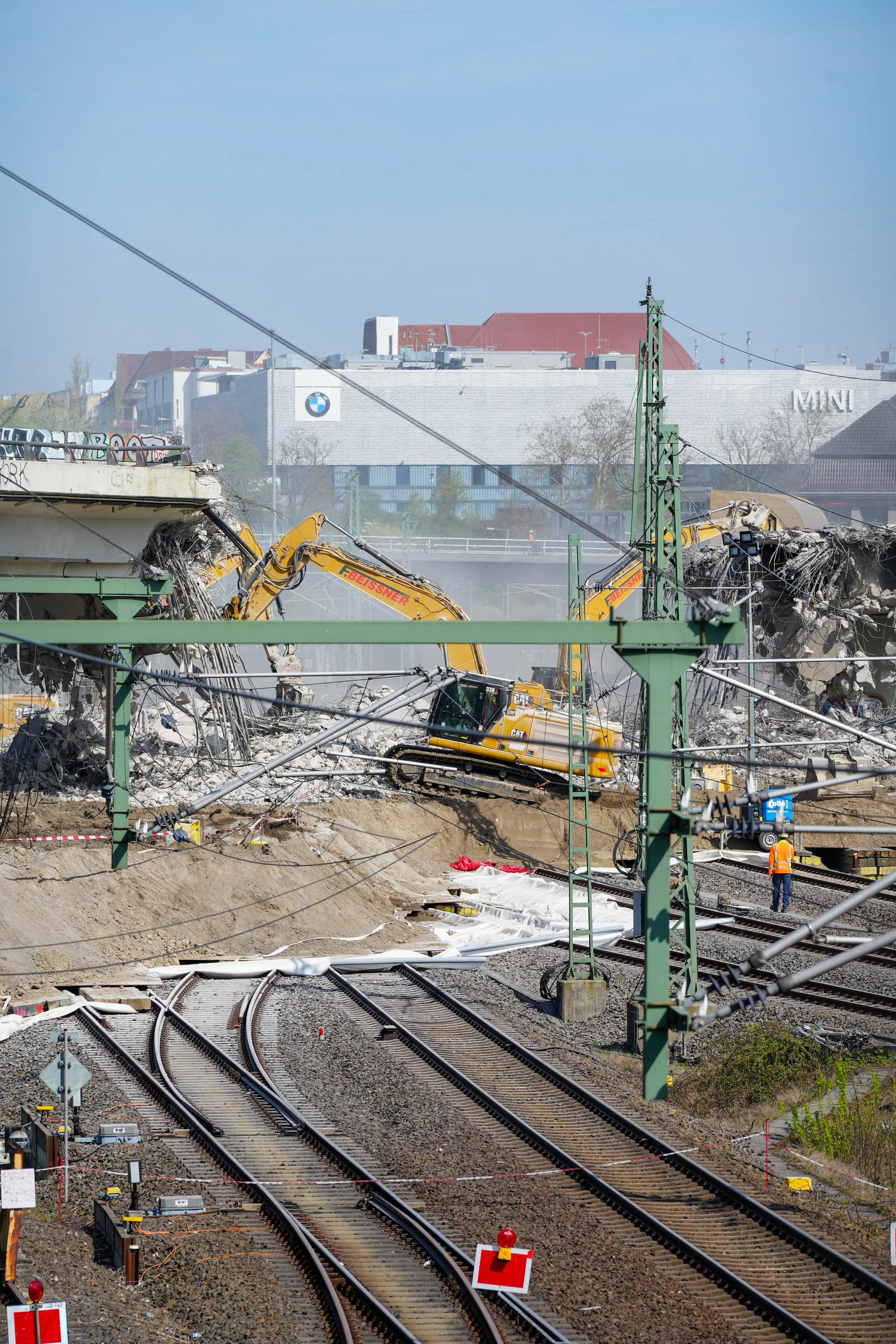 Abbruch einer Brücke über Bahngleisen, mit gelben Baggern und einem Bauarbeiter. Im Hintergrund ein Gebäude mit BMW-Logo.