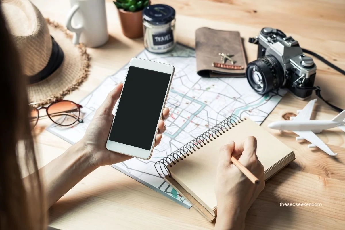 Woman using smartphone and notebook to plan a cruise with maps, travel accessories, and camera gear on the desk.